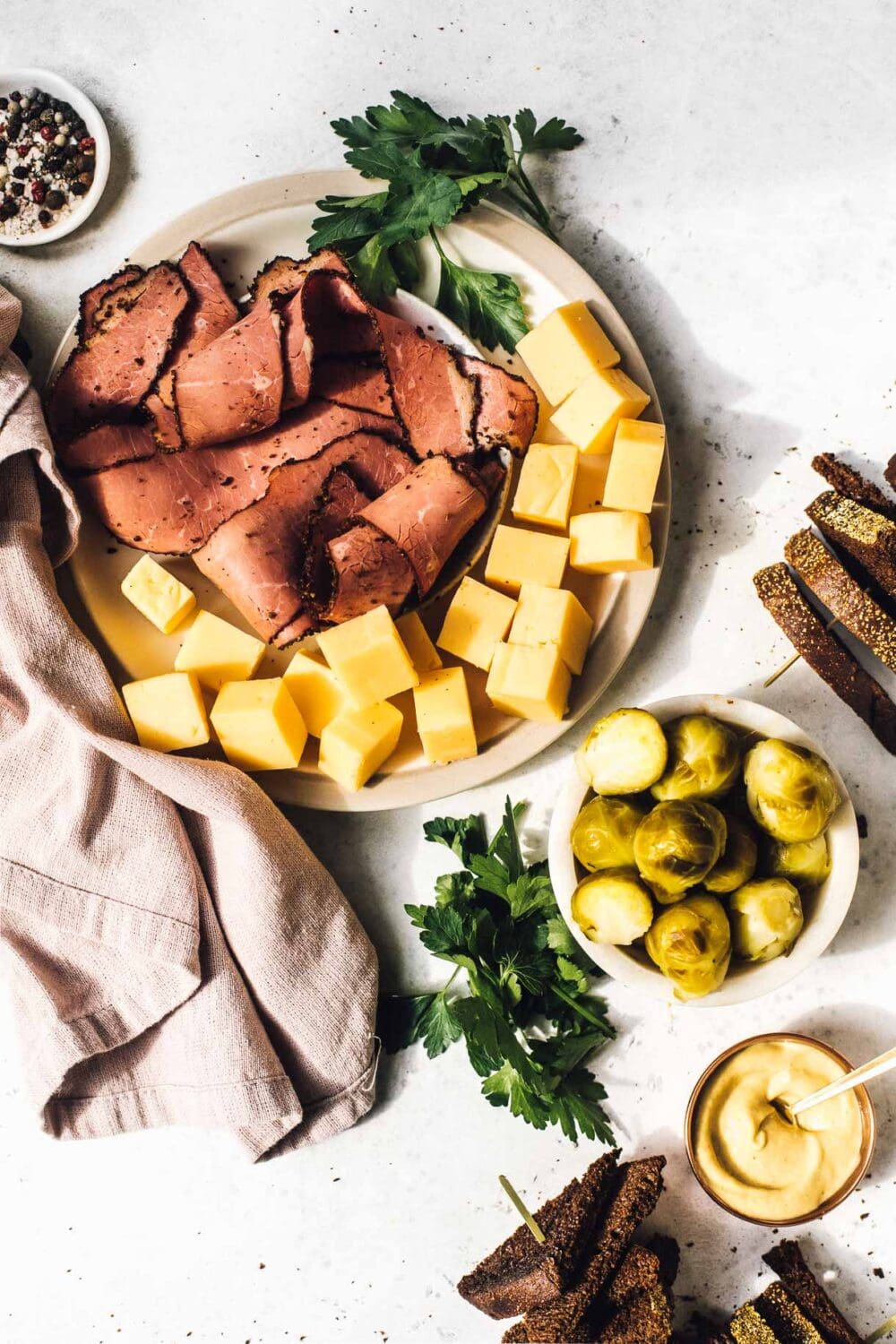 pastrami, cheese, on a white plate with a small plate of pickled brussels sprouts and rye bread next to it.