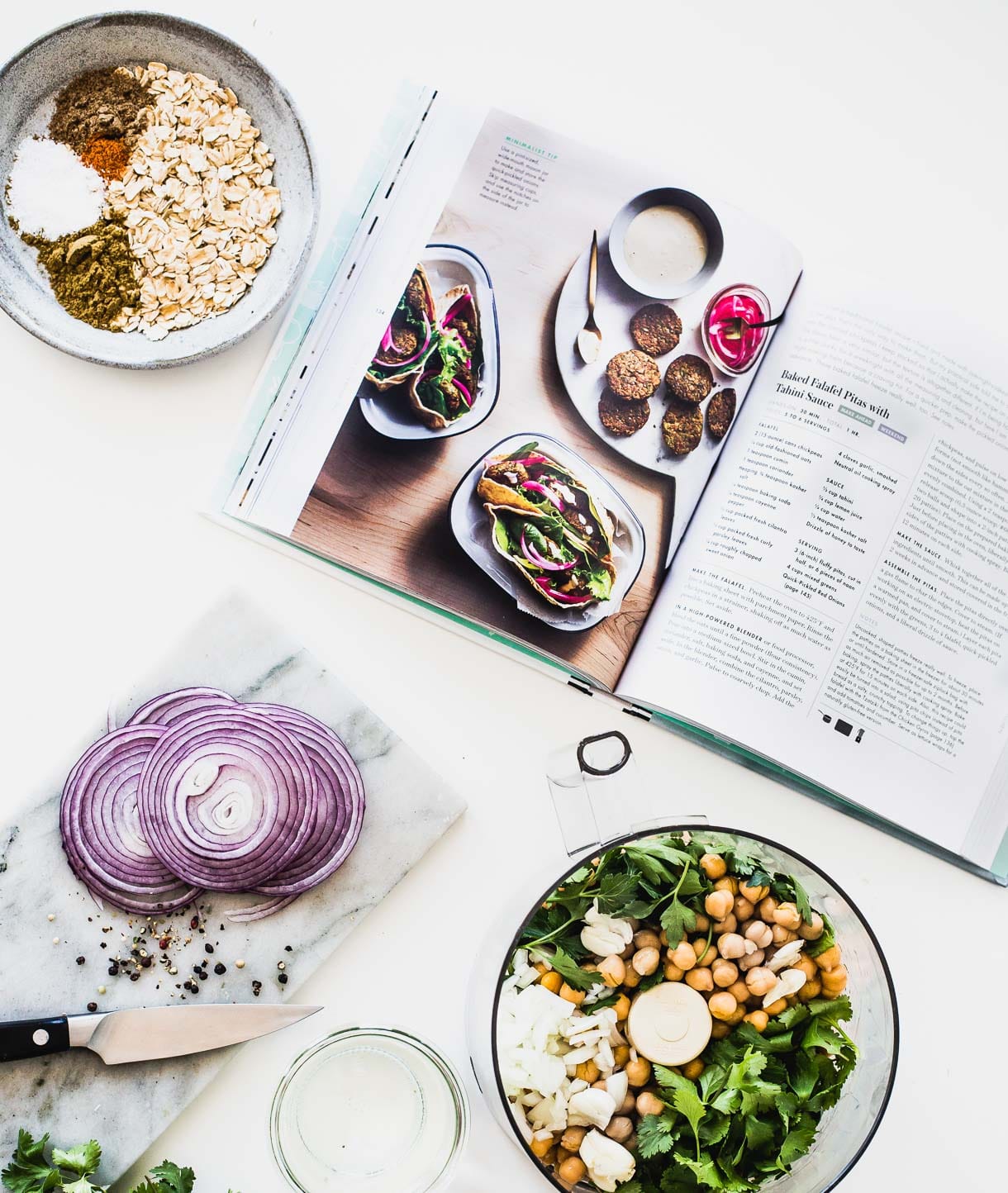 cookbook open, surround by a food processor bowl with ingredients for falafels inside, and cutting board with sliced red onions to the left of it.