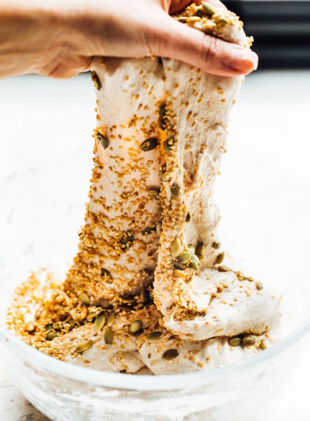 woman performing stretch and fold of seeded sourdough. dough is in a glass bowl.